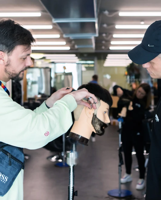 A barbering instructor demonstrating a haircut on a mannequin head while a student in a black cap observes. A barbering instructor demonstrating a haircut on a mannequin head while a student in a black cap observes.